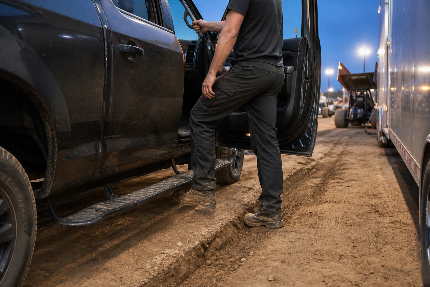 Sprint car crew member tests a pickup running board and grab handle beside a race trailer on a dirt pit lane